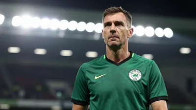 A mid-30s male coach in a dark green Derry City kit, waist-up, intense expression, background blurred stadium lights