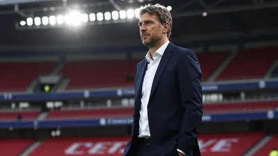 A 34-year-old male football manager in a dark blue suit and white shirt, standing on the sideline at a stadium, focused...