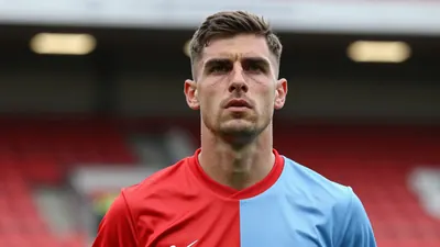 Mid-30s muscular Derry City midfielder, determined expression, head-and-shoulders, wearing a plain red and sky-blue...