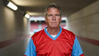 A 50-year-old man with a rugged, weathered face and graying temples, dressed in a plain red and sky blue football jersey...
