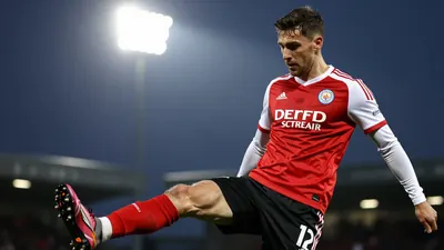 A tight waist-up shot of a Derry City player in a red jersey with white sleeves and black shorts, looking focused as they...