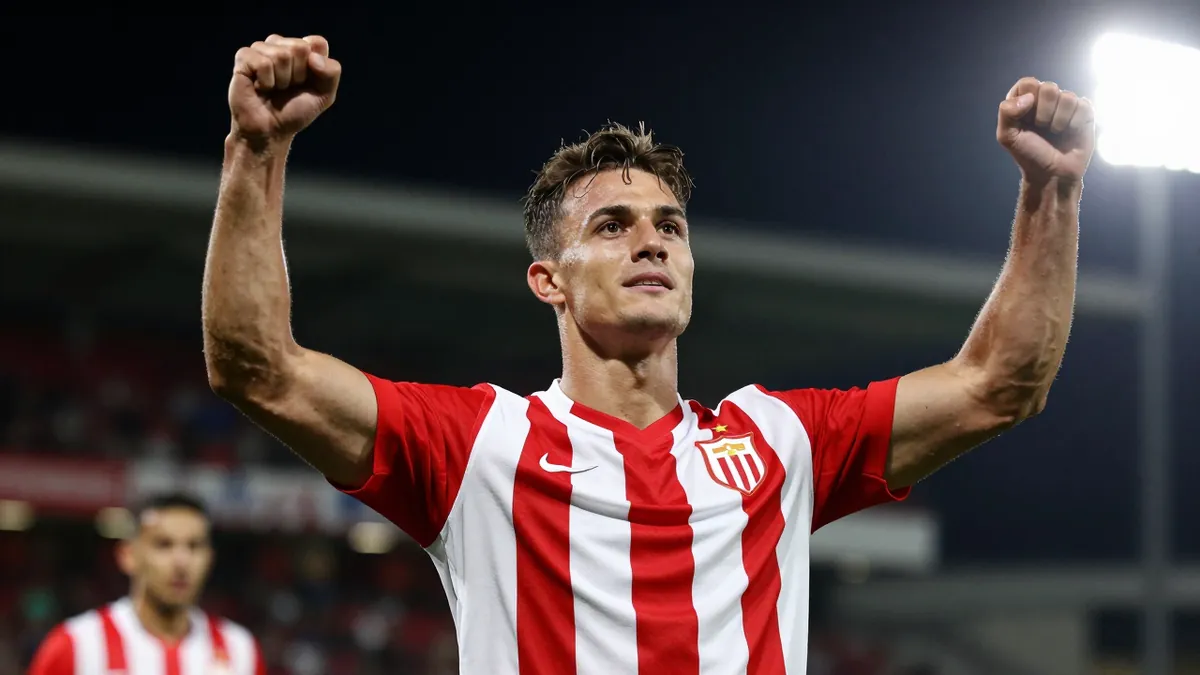 A muscular young male footballer in a red and white striped shirt, celebrating a goal with arms raised